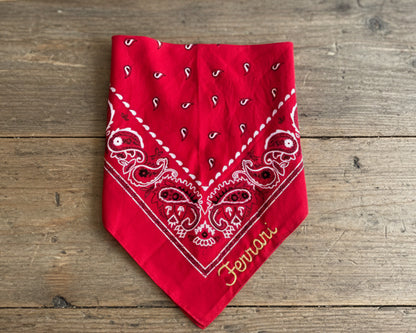 A high-performance red paisley bandana featuring the "Ferrari" name in premium gold chain stitch embroidery, displayed on a rustic wooden background.