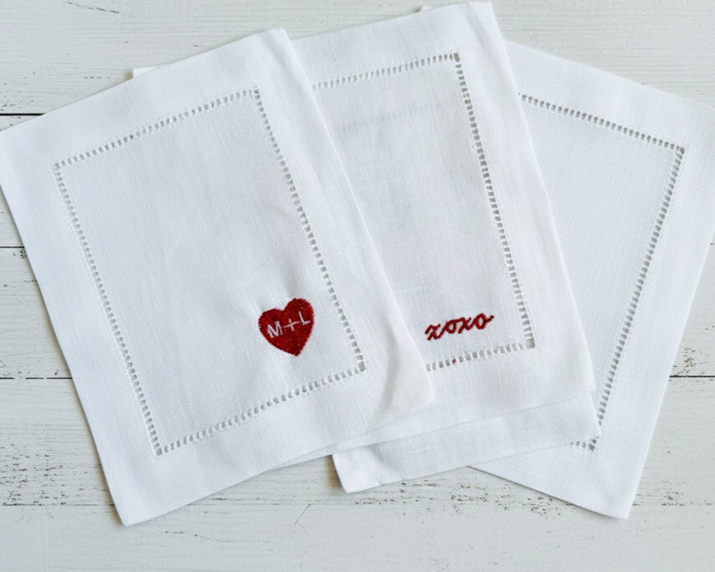 Three white napkins with embroidered designs on a light wooden surface