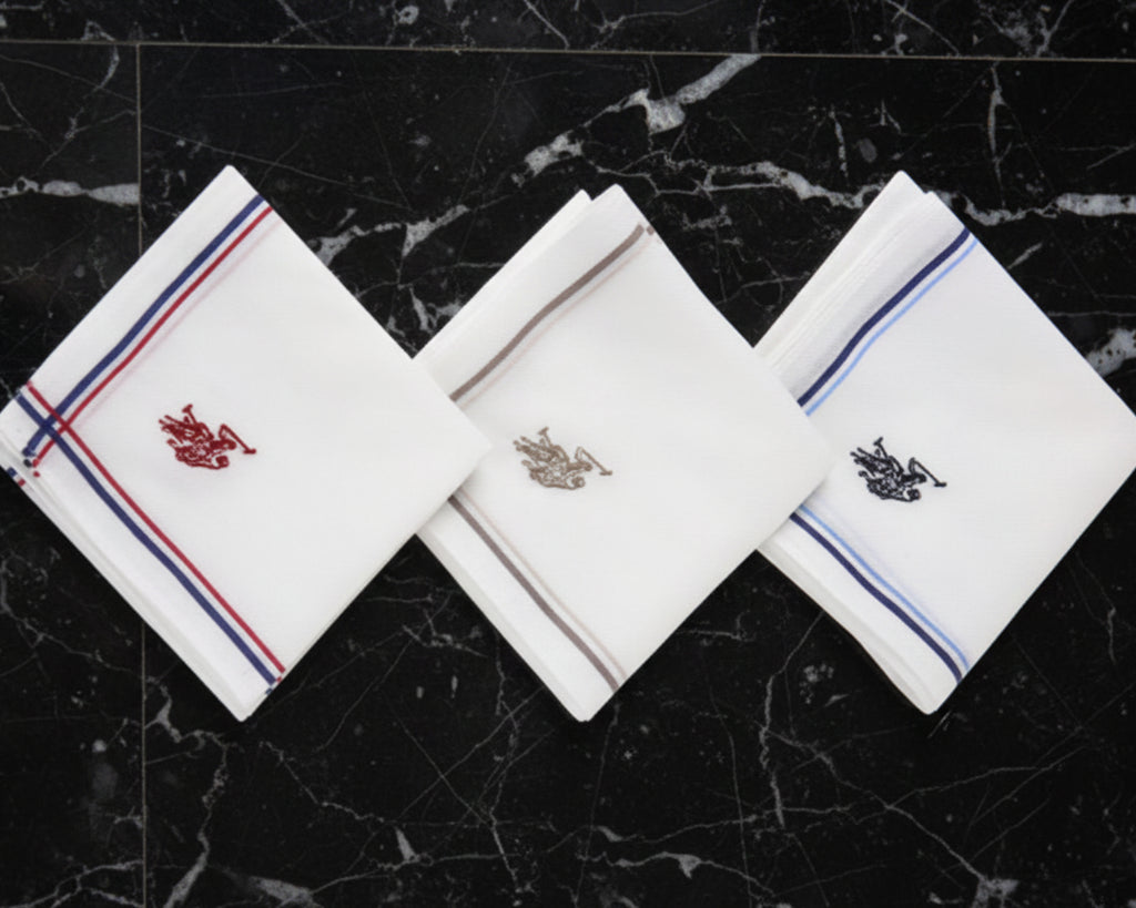 A white cotton handkerchief by U.S. Polo Assn. with gold striped borders and a royal blue embroidered brand logo.