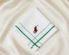 A crisp white U.S. Polo Assn. cotton pocket square featuring green striped borders and a red embroidered horseman logo.