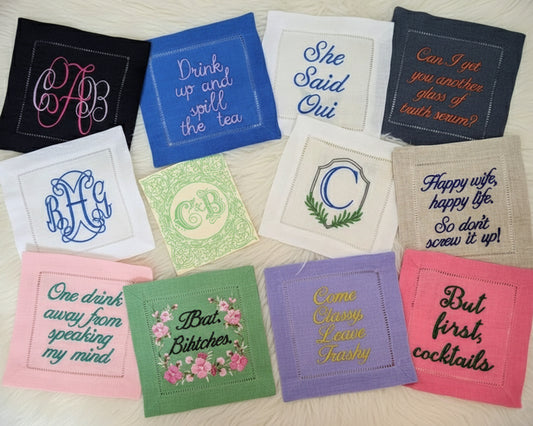 A group of colorful scalloped-edge linen napkins in rose pink, sky blue, and white featuring custom bar name embroidery and cocktail icons.