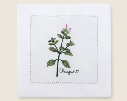 Single white linen cocktail napkin featuring a detailed purple-flowered Cardamom plant embroidery with a hemstitch border.