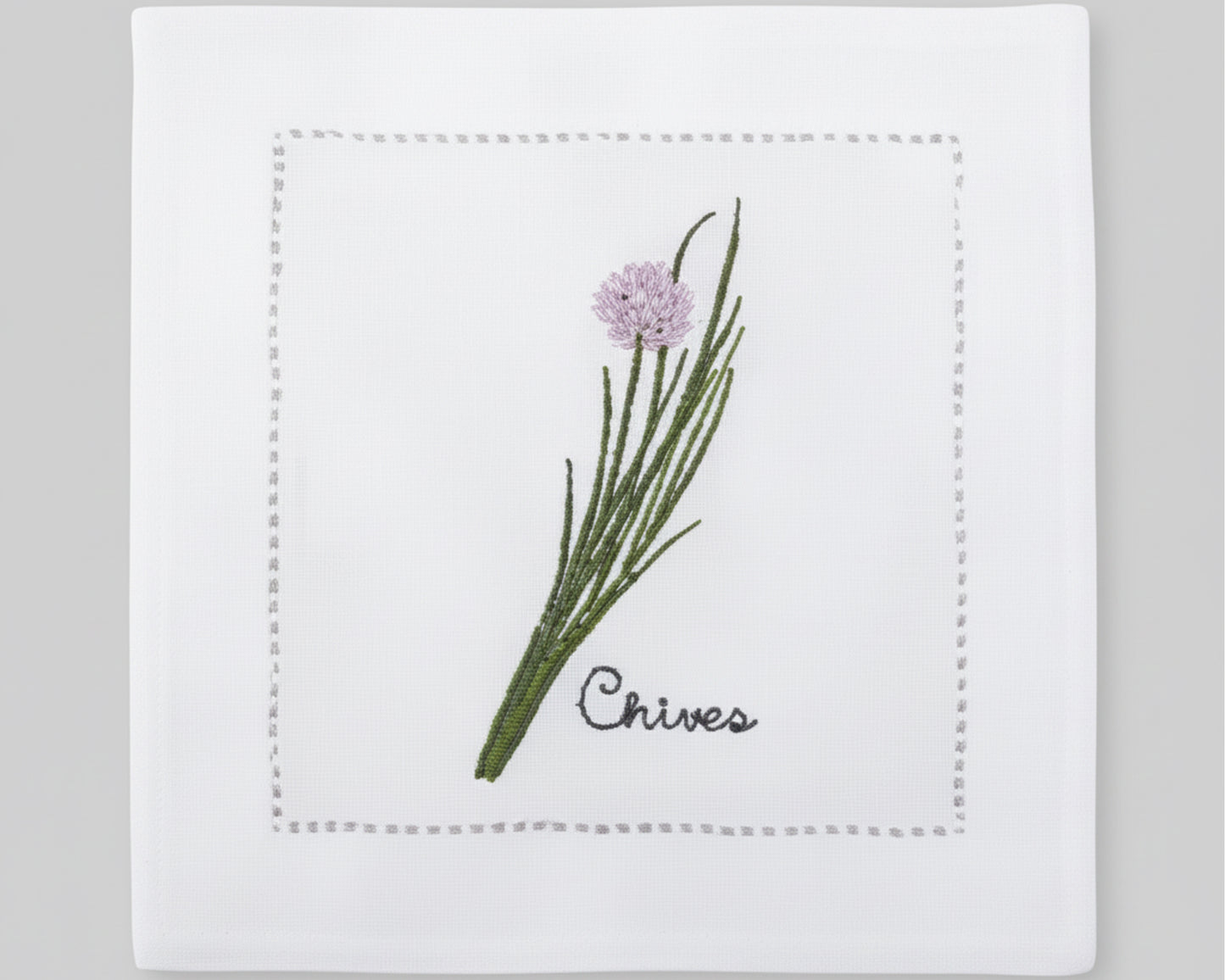 Single white linen cocktail napkin featuring a detailed purple-flowered Chive embroidery with a delicate hemstitch border.