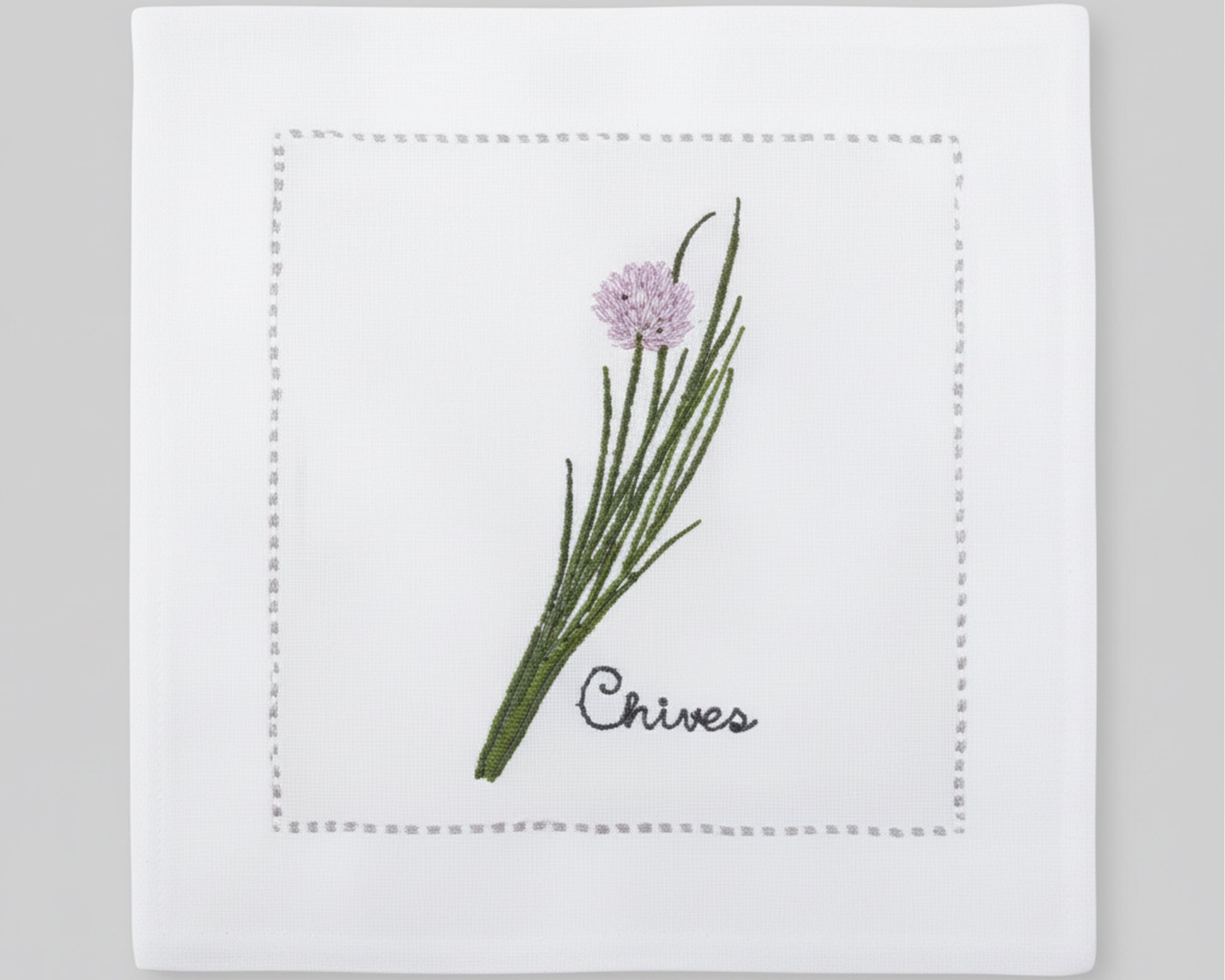 Single white linen cocktail napkin featuring a detailed purple-flowered Chive embroidery with a delicate hemstitch border.