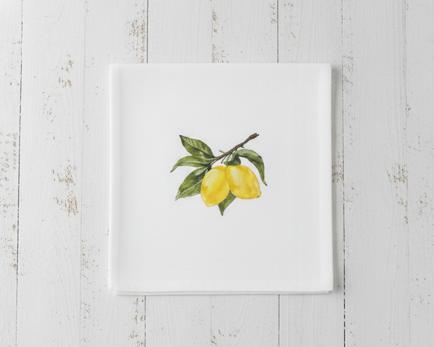 Close up of a single white linen cocktail napkin featuring a sun-ripened yellow lemon botanical embroidery centered on a crisp white fabric.