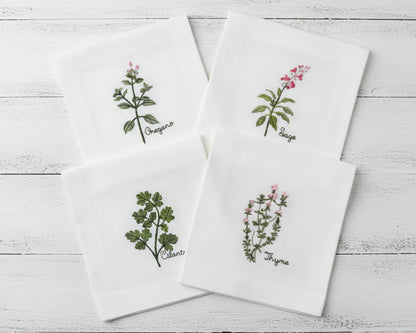 A set of four white linen cocktail napkins featuring embroidered Oregano, Sage, Cilantro, and Thyme botanical designs.