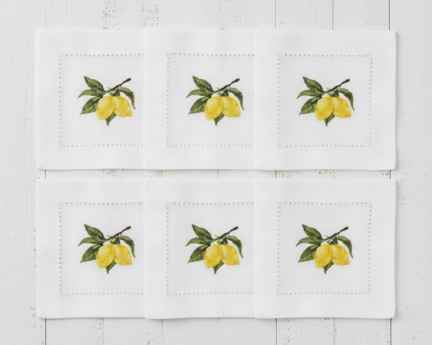 A set of six white linen cocktail napkins each featuring a detailed yellow lemon and green leaf branch embroidery with a hemstitch border.