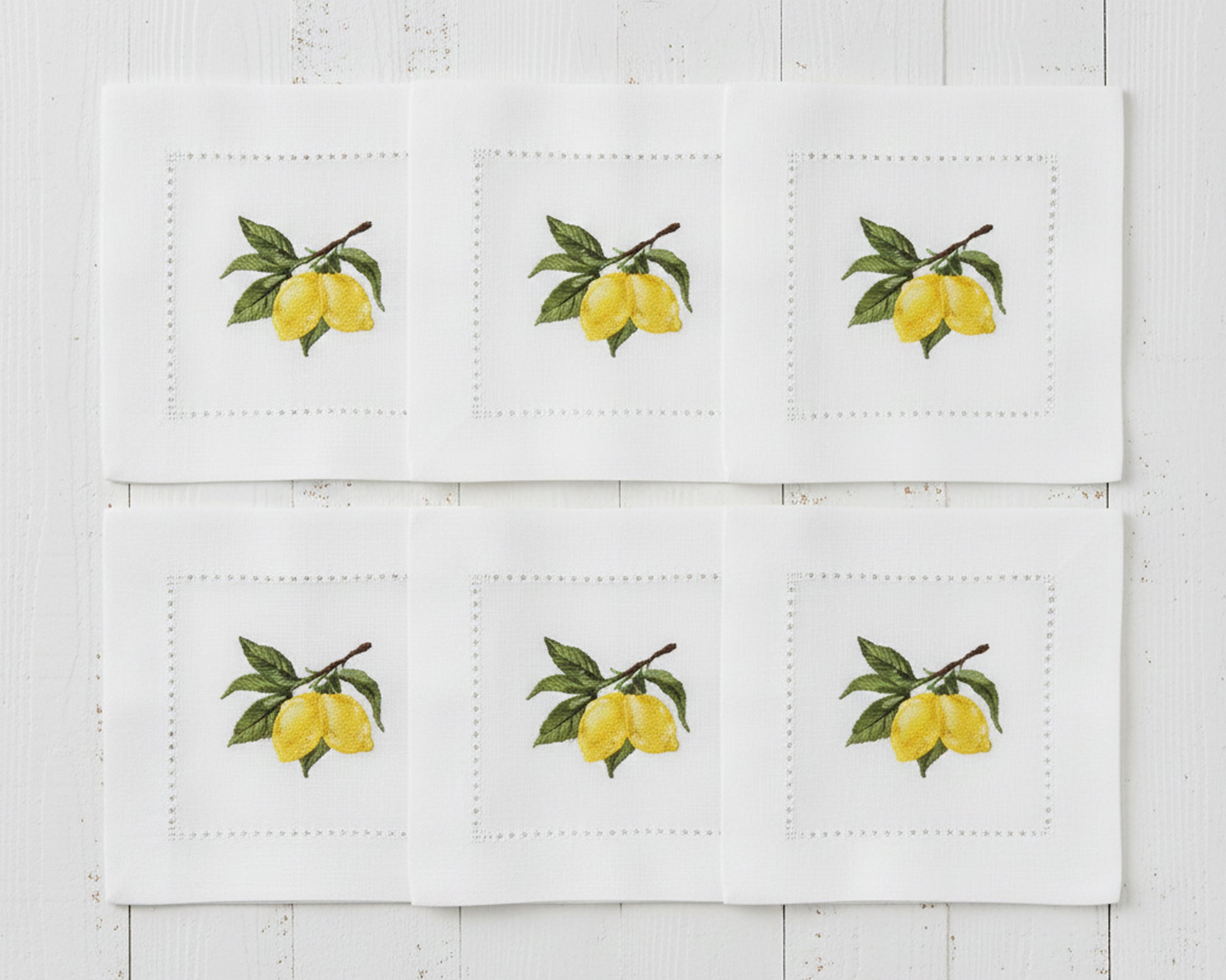 A set of six white linen cocktail napkins each featuring a detailed yellow lemon and green leaf branch embroidery with a hemstitch border.