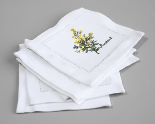 Close up of a stacked white linen napkin featuring a detailed yellow Mustard flower embroidery with green leaves.
