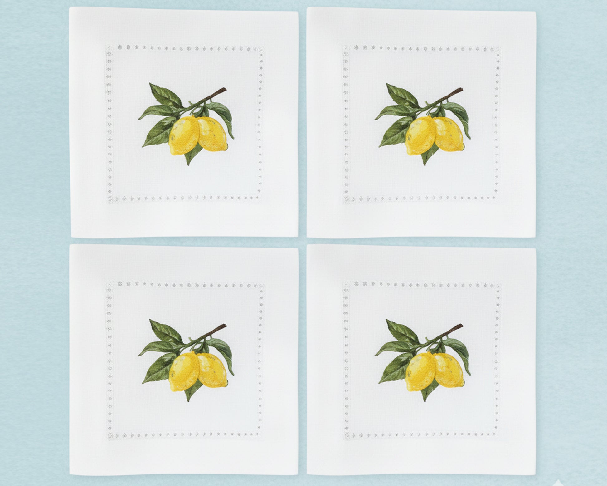 A set of six pure white cotton cocktail napkins featuring detailed yellow lemon and green leaf branch embroidery with a hemstitch border.