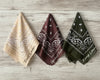 Three folded paisley bandanas in tan, brown, and olive with custom phrases like "Treat Lover"