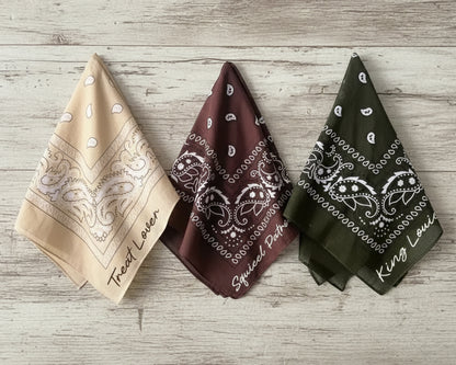Three folded paisley bandanas in tan, brown, and olive with custom phrases like "Treat Lover"