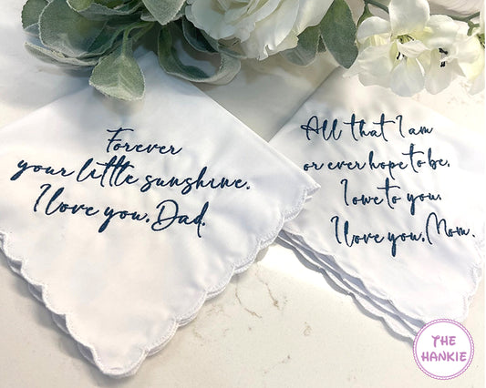 Two white handkerchiefs with embroidered messages on a marble surface with flowers.