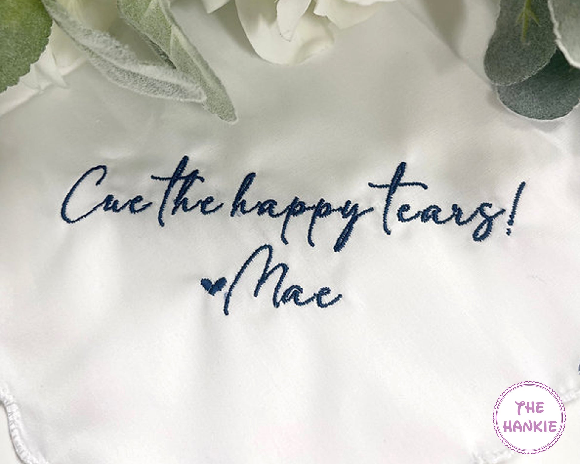 White hankie with embroidered text ' Cue the happy tears! Mae' on a white background with greenery.