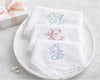 Set of white embroidered napkins with initials on a white plate, gift box, and earrings in the background.