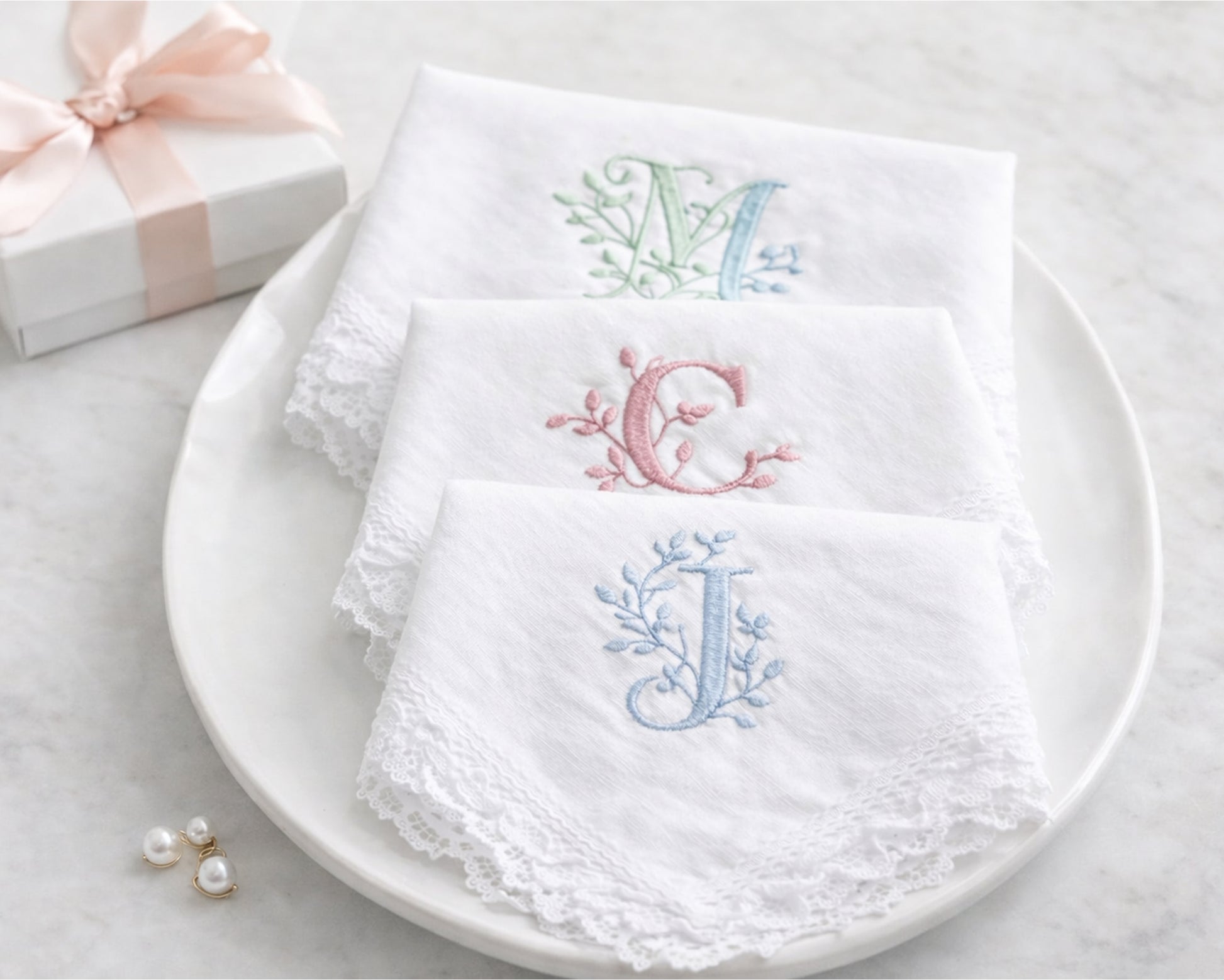 Set of white embroidered napkins with initials on a white plate, gift box, and earrings in the background.