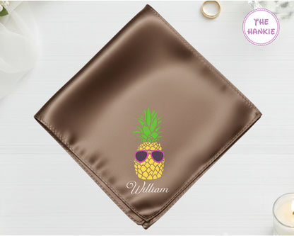 A burnished bronze satin handkerchief featuring "William" and a detailed pineapple embroidery wearing pink-lensed sunglasses.